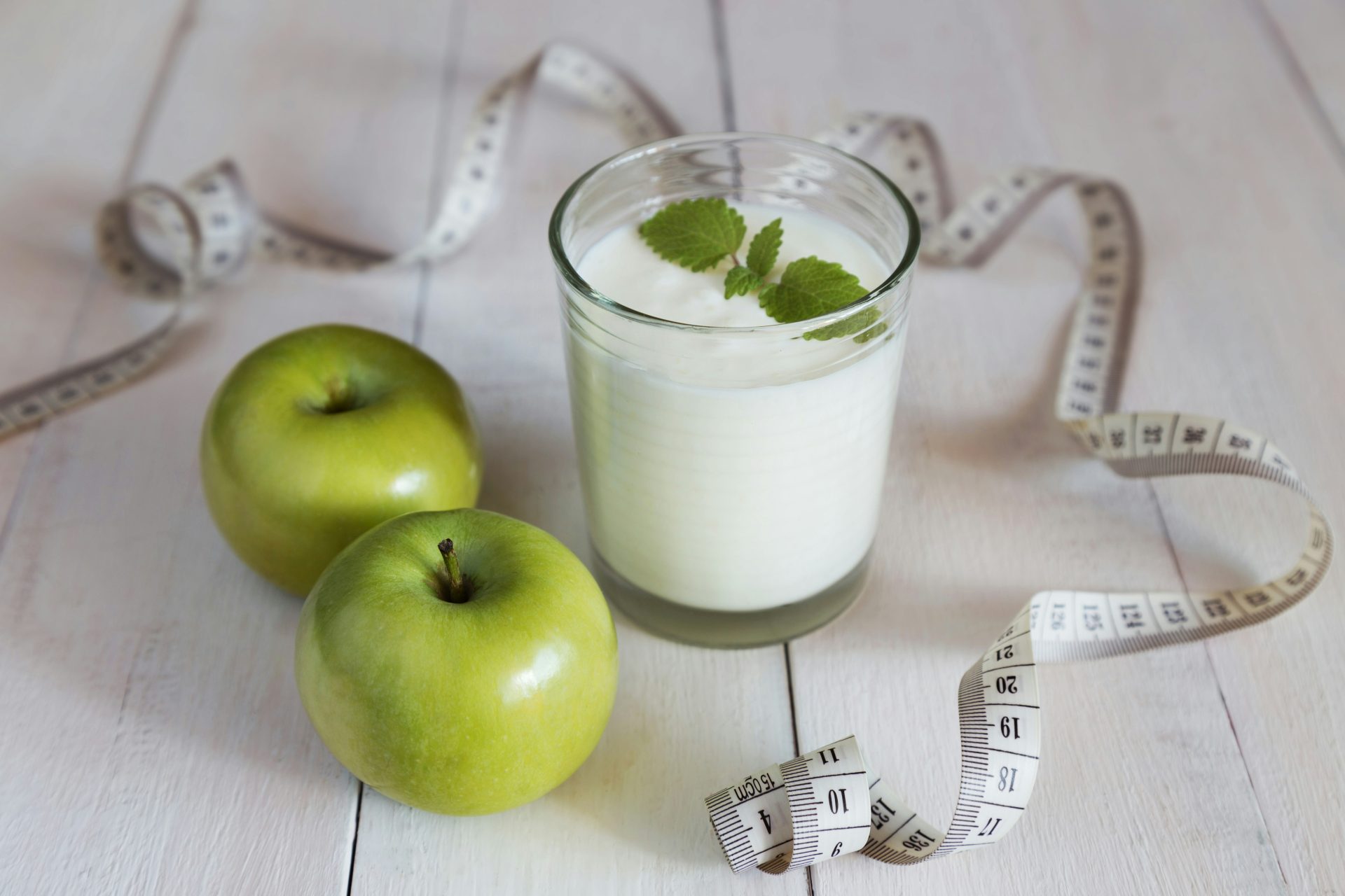 Un vaso de leche junto a dos manzanas verdes y una cinta métrica.